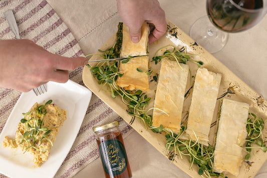 Main coupant un feuilleté aux épinards, à côté d'une assiette de tartinade de pois chiches, une coupe de vin rouge ainsi qu'une bouteille de vinaigres aromatisés. 