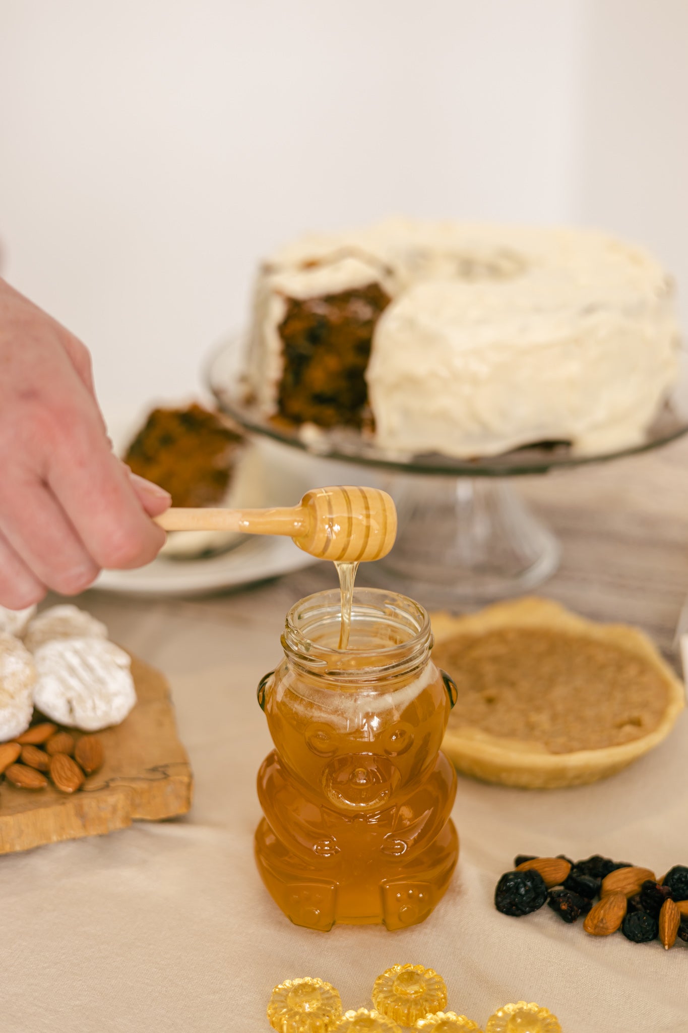 Un pot de miel de fleurs sauvages en forme d'ourson déposé sur une table à coté de desserts tel qu'une tarte sucré et un gâteau.